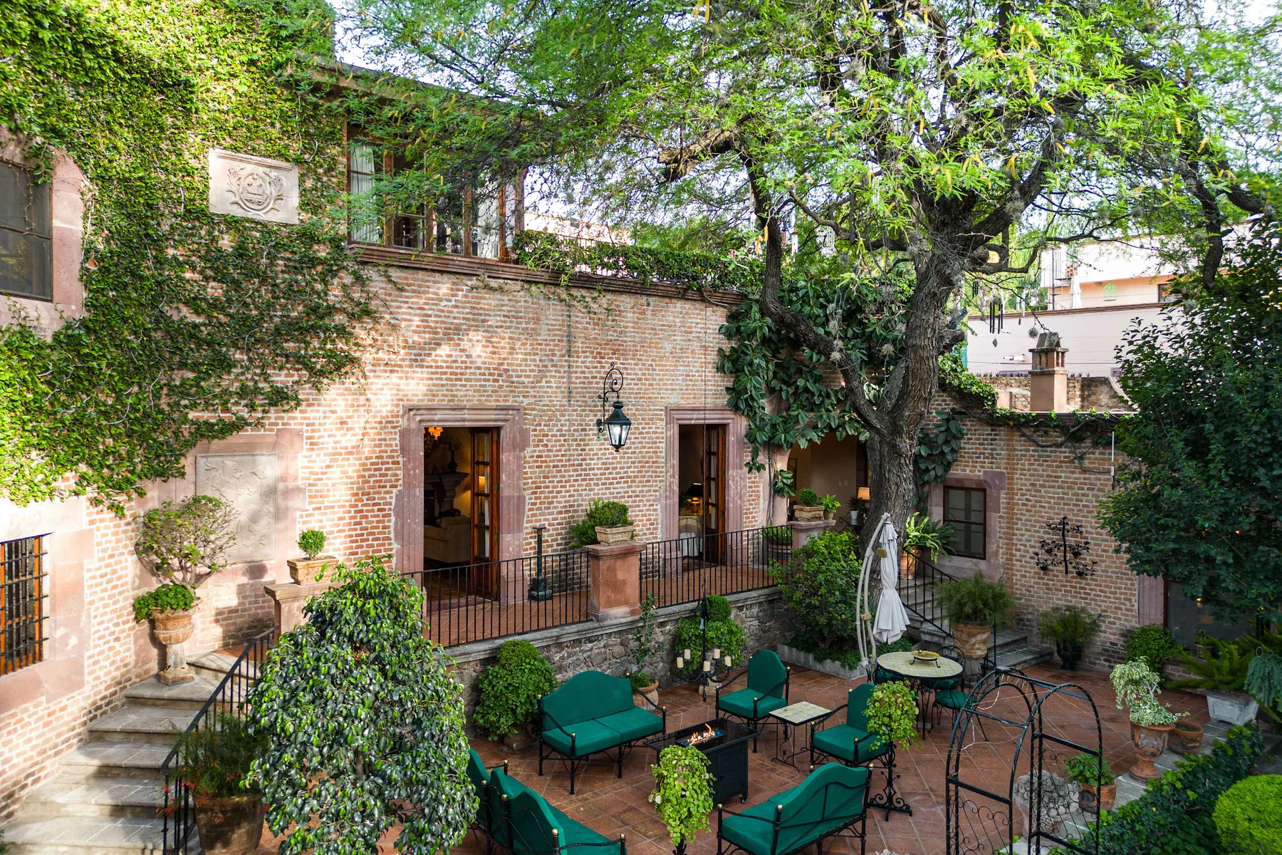 A brick courtyard patio has green seating beneath a leafy tree.
