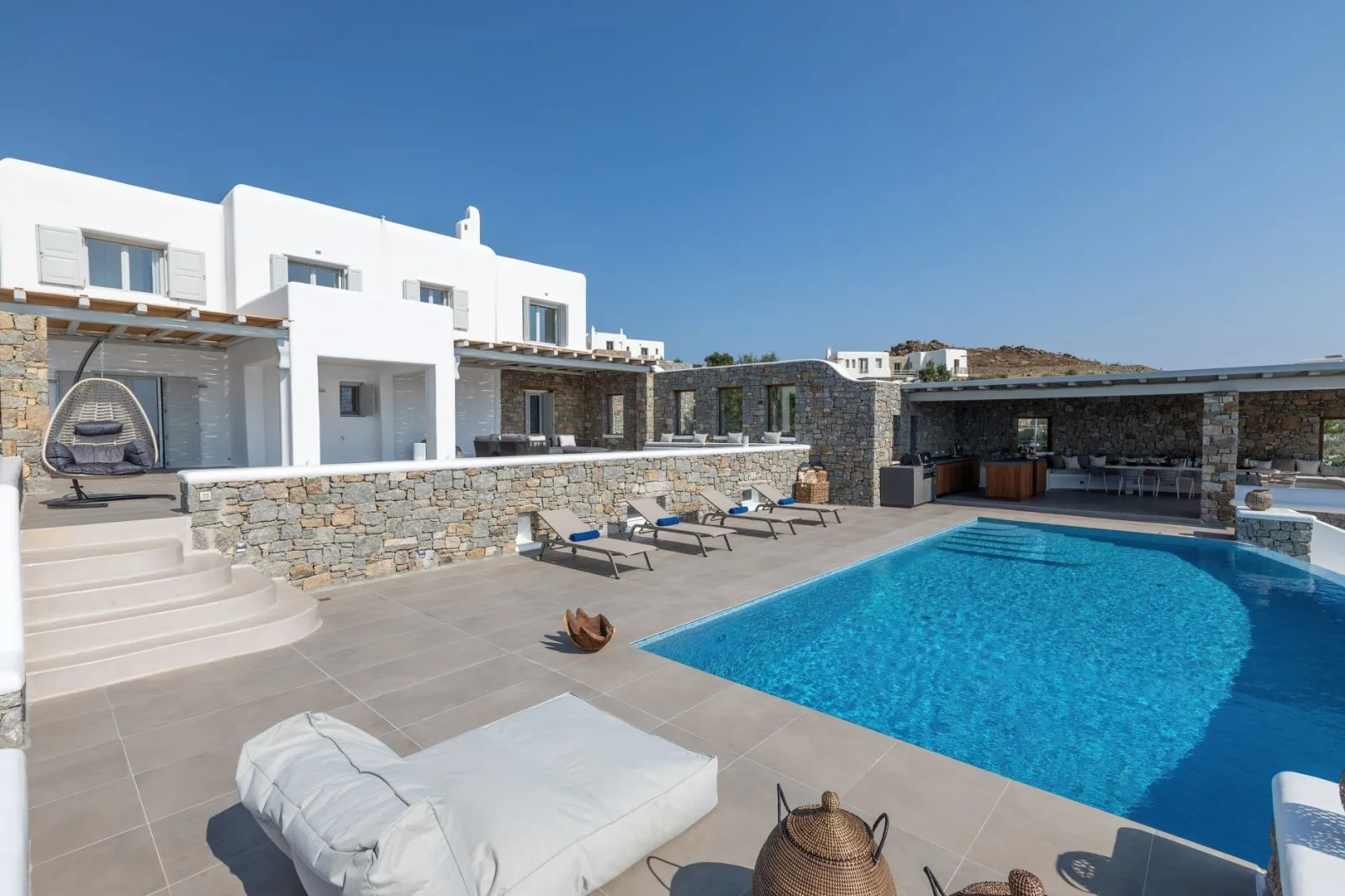 A sunlit pool terrace beside a white building, with lounge seating and a stone wall.