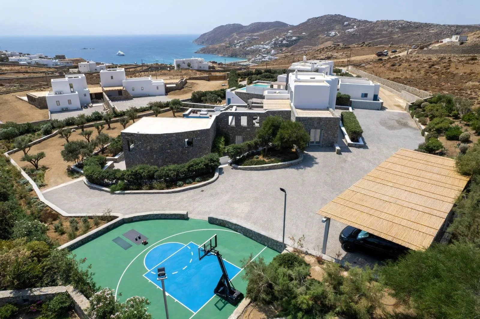An outdoor basketball court sits beside a low stone building, with the sea in the distance.