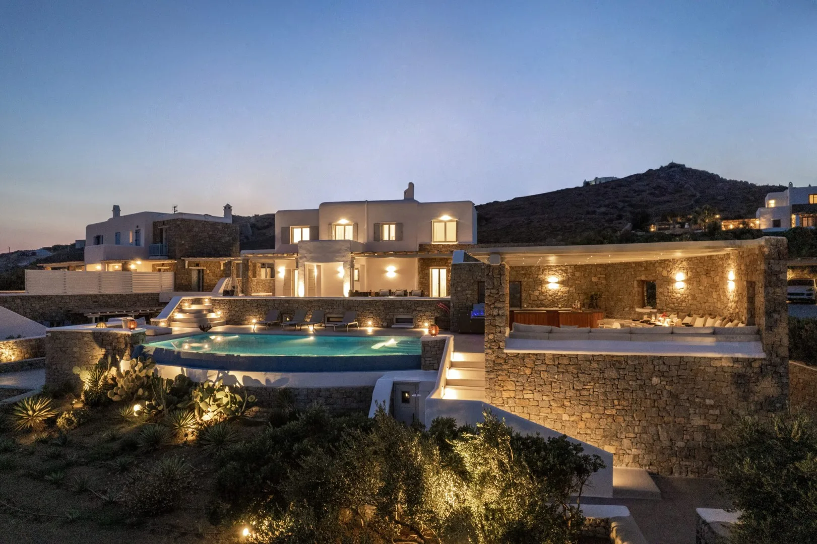A modern house and illuminated swimming pool sit against a dark hillside at dusk.