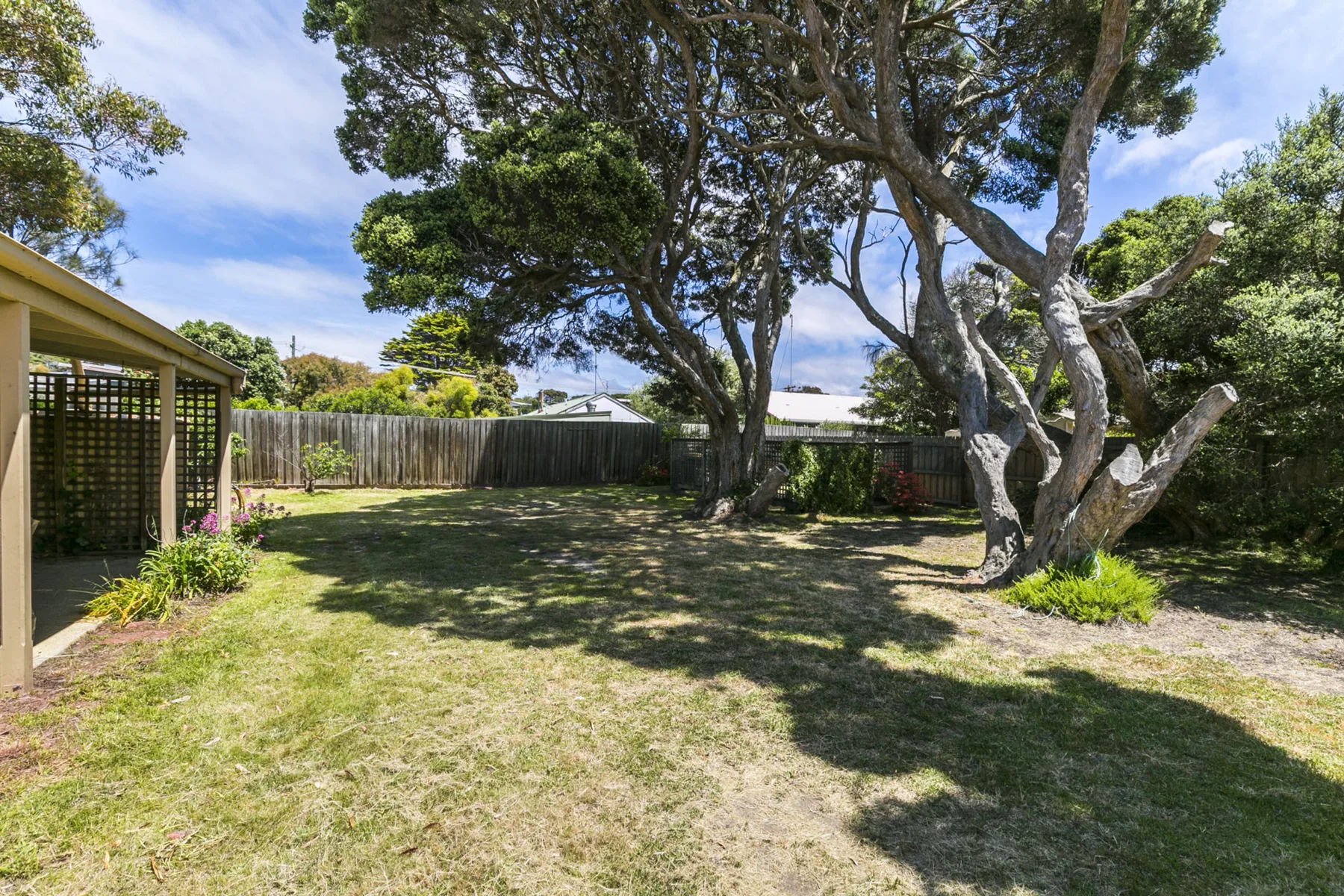 A grassy yard with mature trees, a wooden fence, and a covered patio.