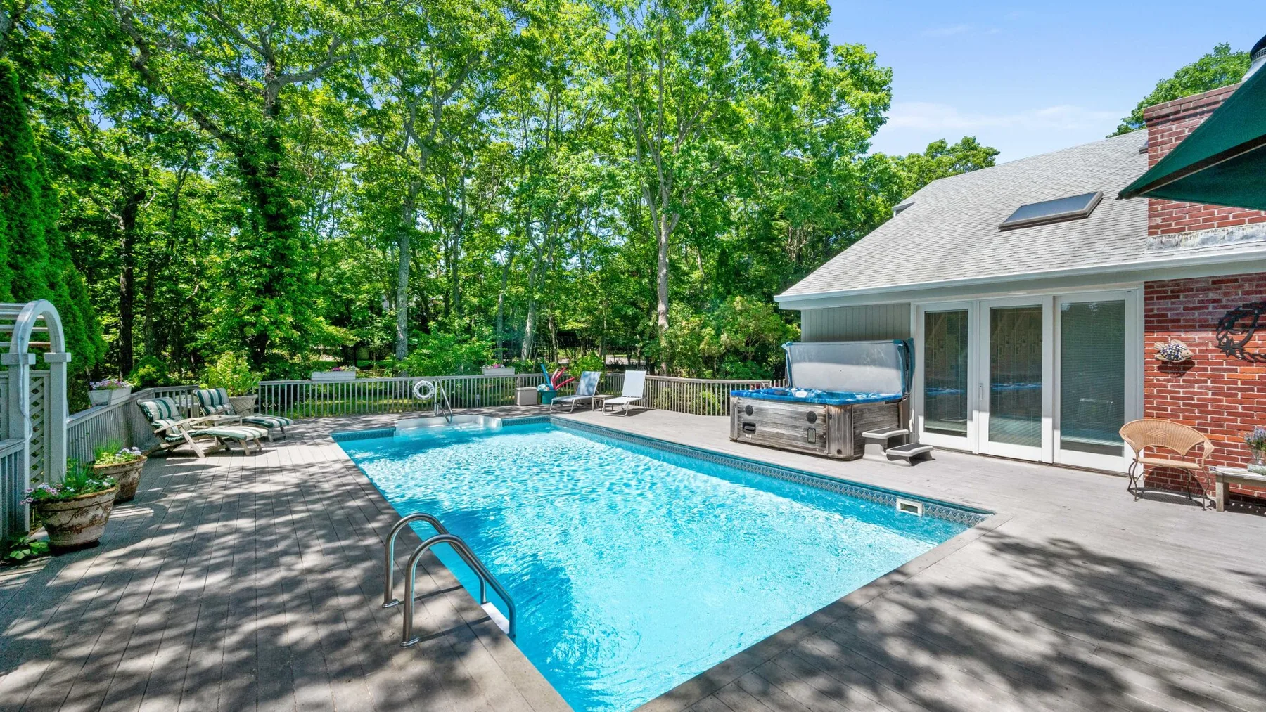 An outdoor swimming pool sits beside a house, surrounded by a wooded yard.