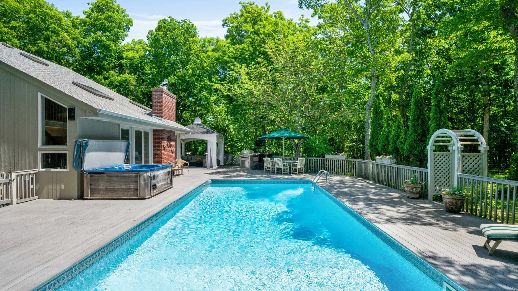 An outdoor swimming pool fills a spacious deck beside a house and leafy trees.