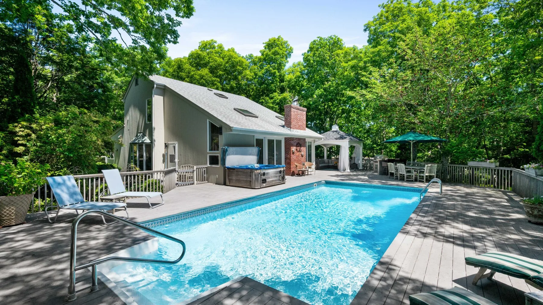 A swimming pool sits beside a house on a sunlit wooden deck surrounded by trees.