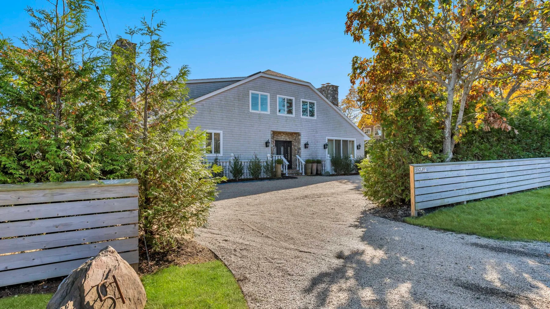 A gray house sits at the end of a gravel driveway bordered by trees and fencing.