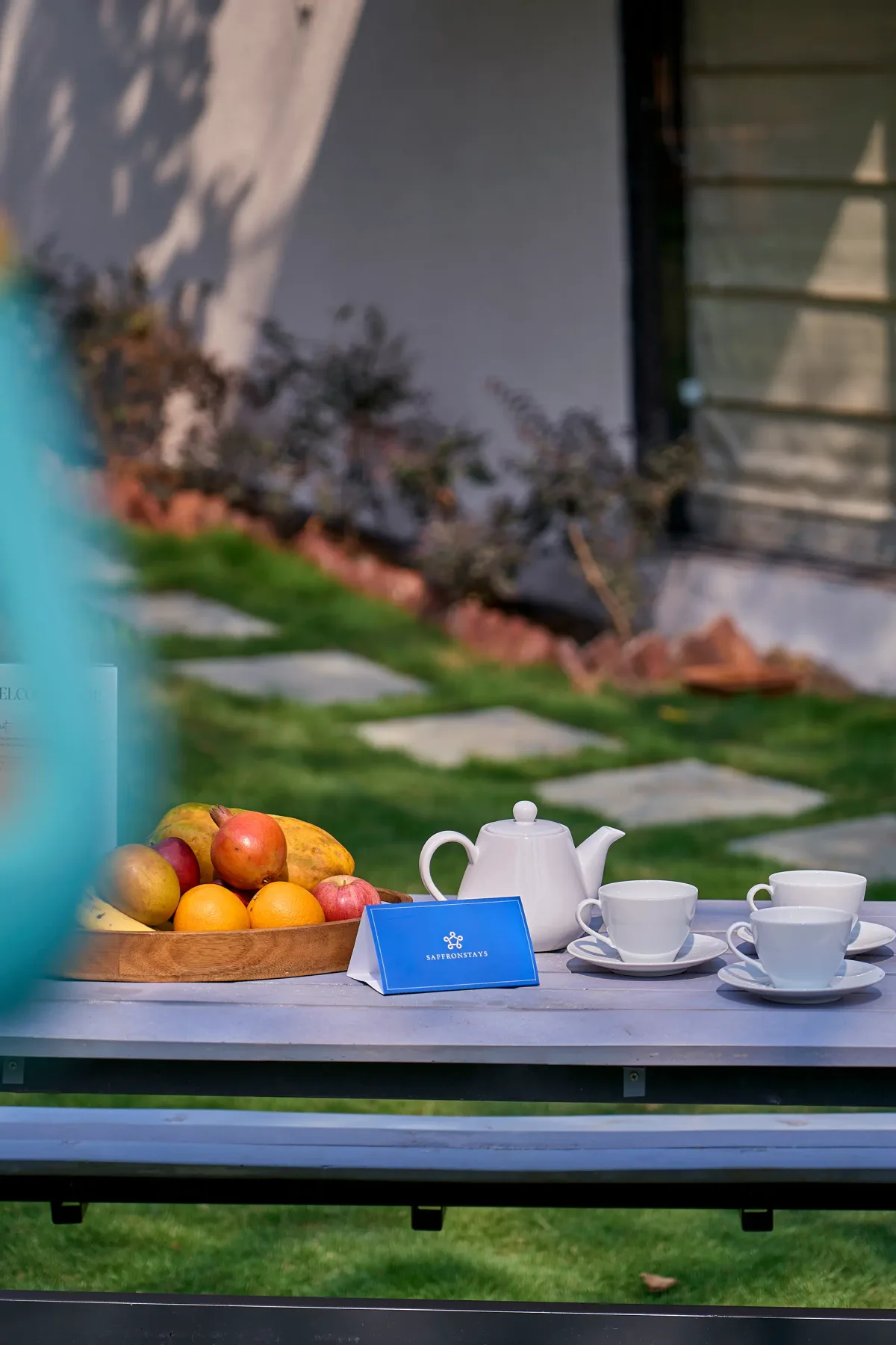 Una mesa de jardín muestra fruta, una tetera, tazas y un pequeño letrero.