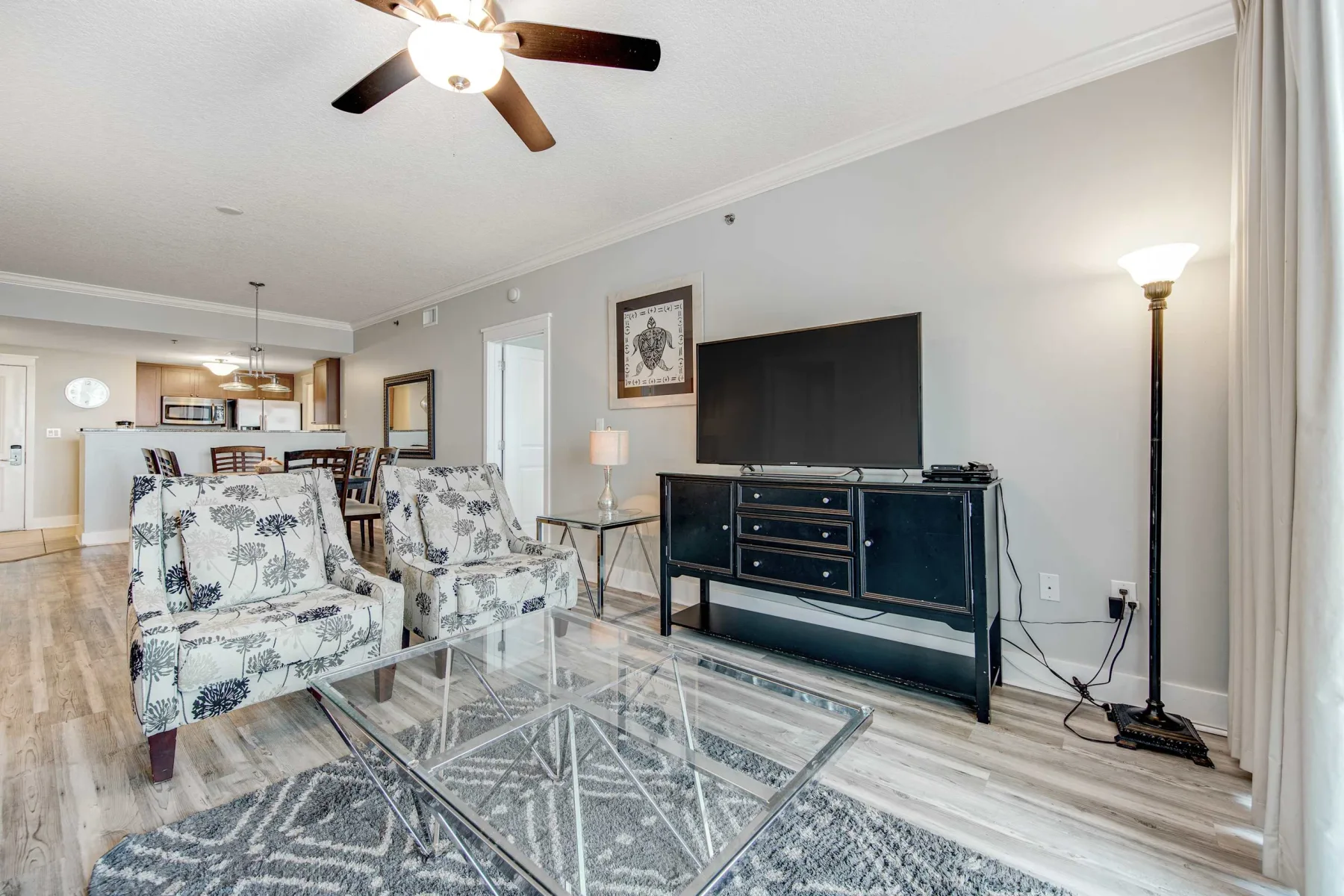 A bright living room with patterned seating, a television, and a glass coffee table.