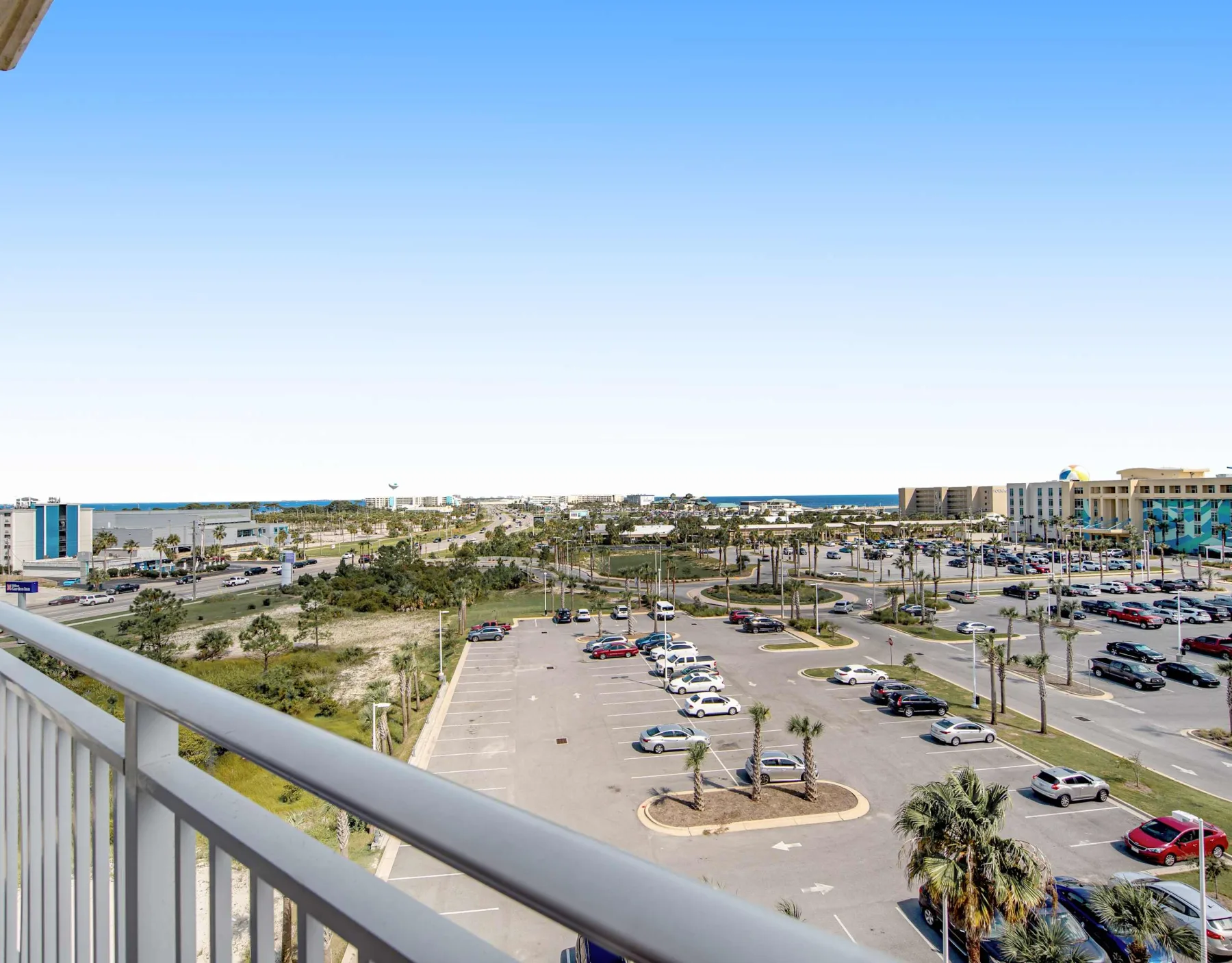 A balcony overlooks a parking lot, palm trees, and a distant strip of coastline.