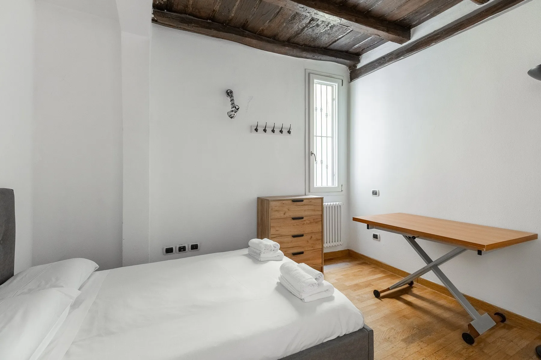 A bright bedroom with a bed, wooden dresser, and desk beneath exposed ceiling beams.