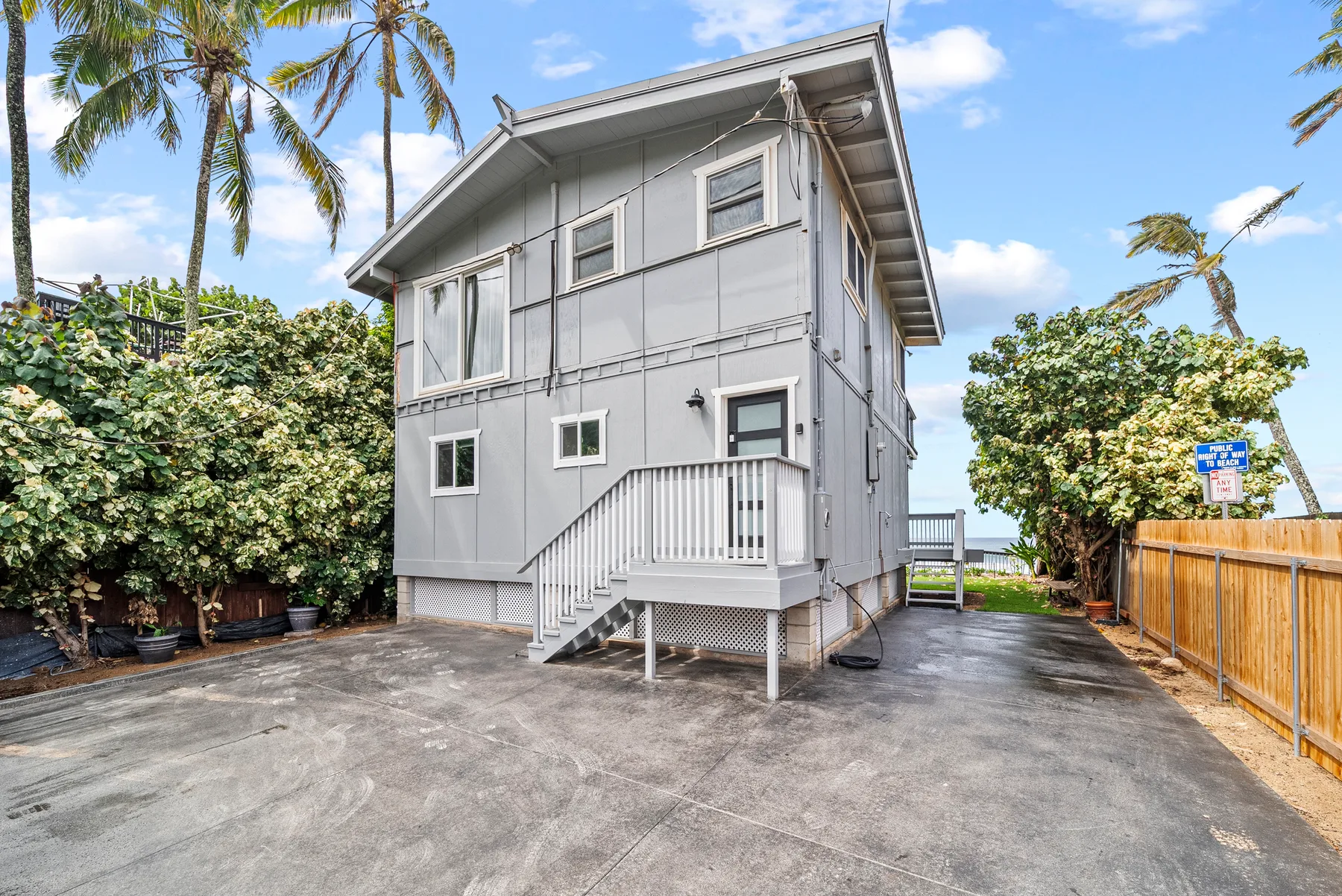 A gray raised house stands beside a paved driveway, with tropical plants and palm trees.