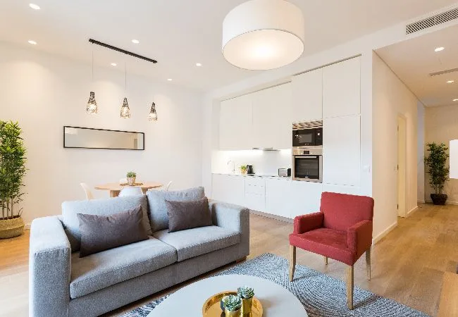 Una luminosa sala de estar con sofá gris, sillón rojo y cocina abierta.