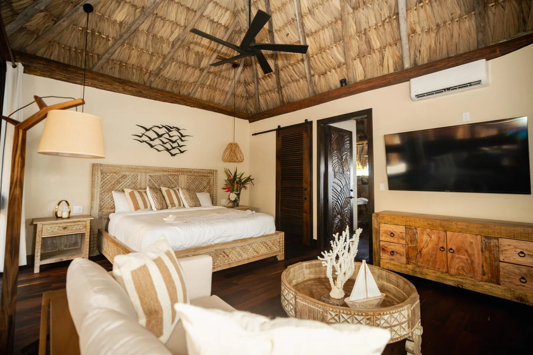 A bedroom with a bed, seating, and a wall-mounted television beneath a thatched ceiling.