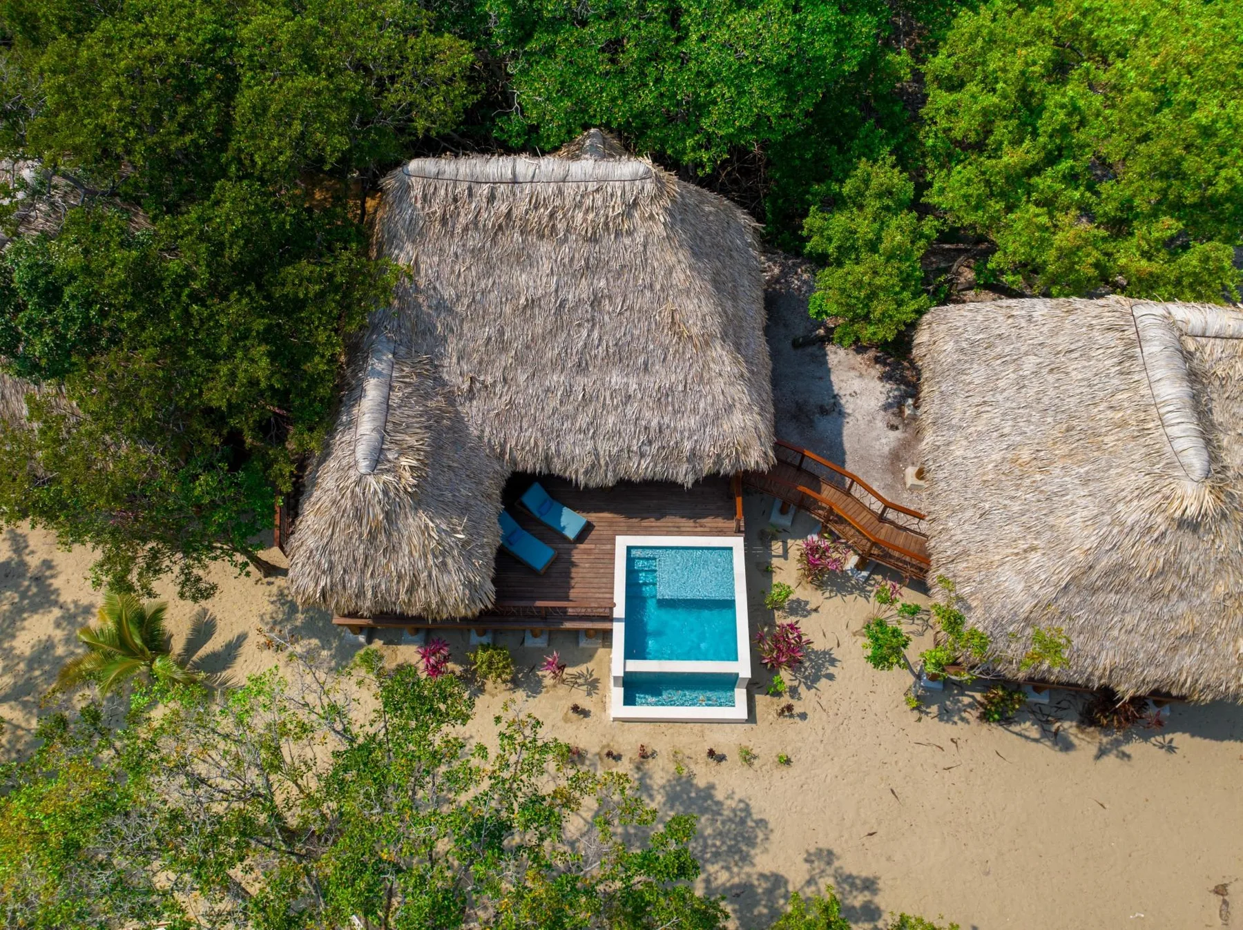 An aerial view of thatched-roof buildings beside a small rectangular pool in a sandy clearing.
