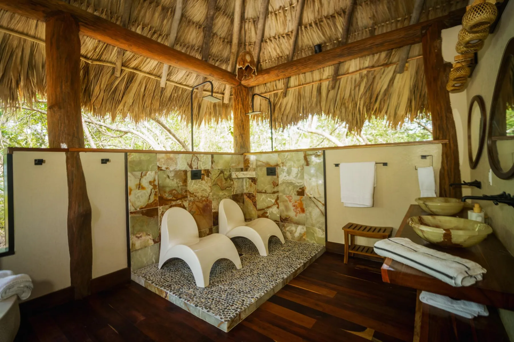 An open-air bathroom with a tiled shower area beneath a thatched roof.