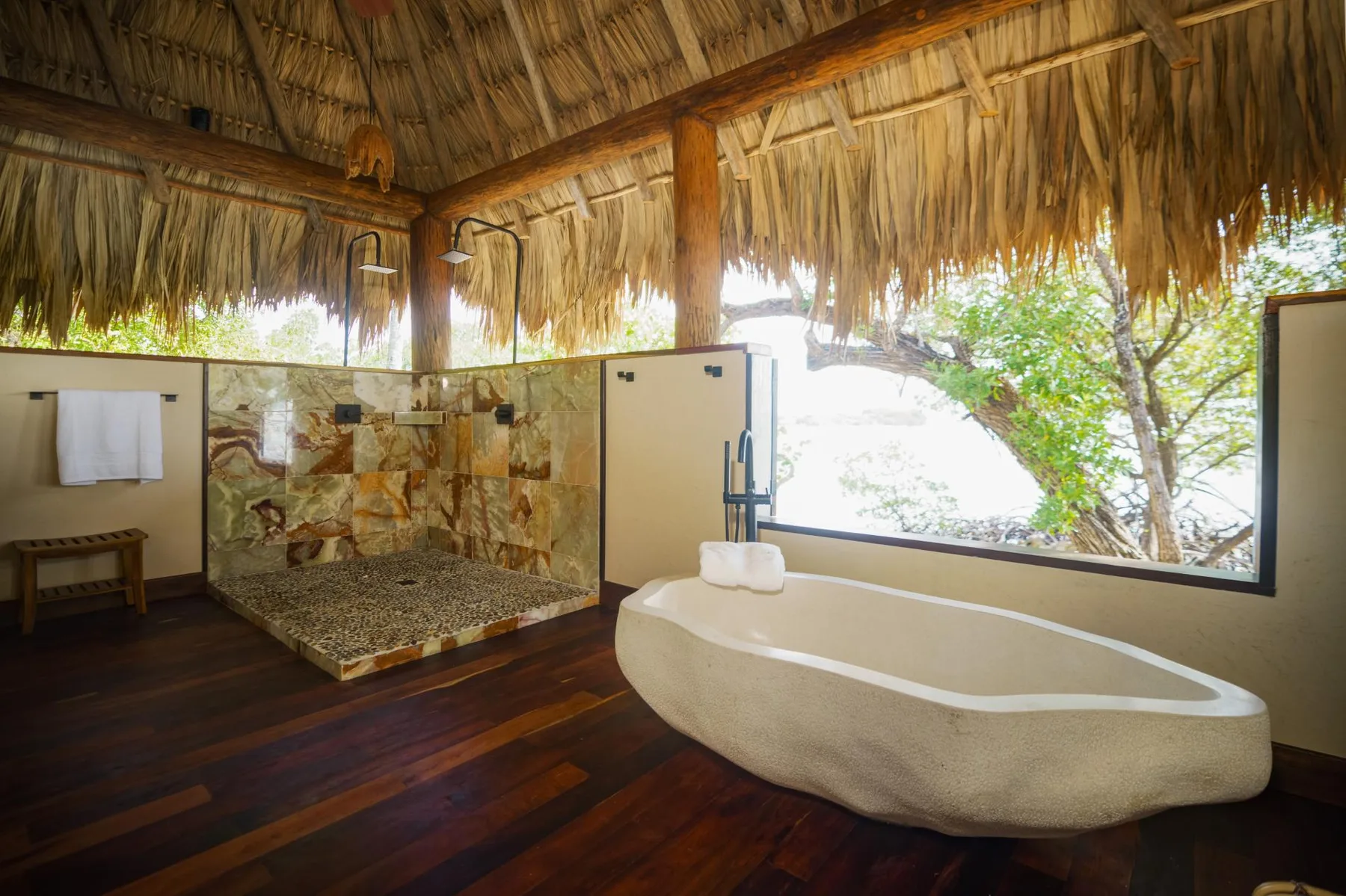 An open-air bathroom with a freestanding bathtub beneath a thatched roof.