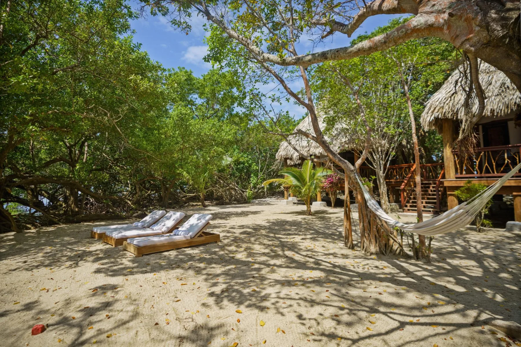 A sandy outdoor seating area sits beneath trees beside thatched huts and a hammock.