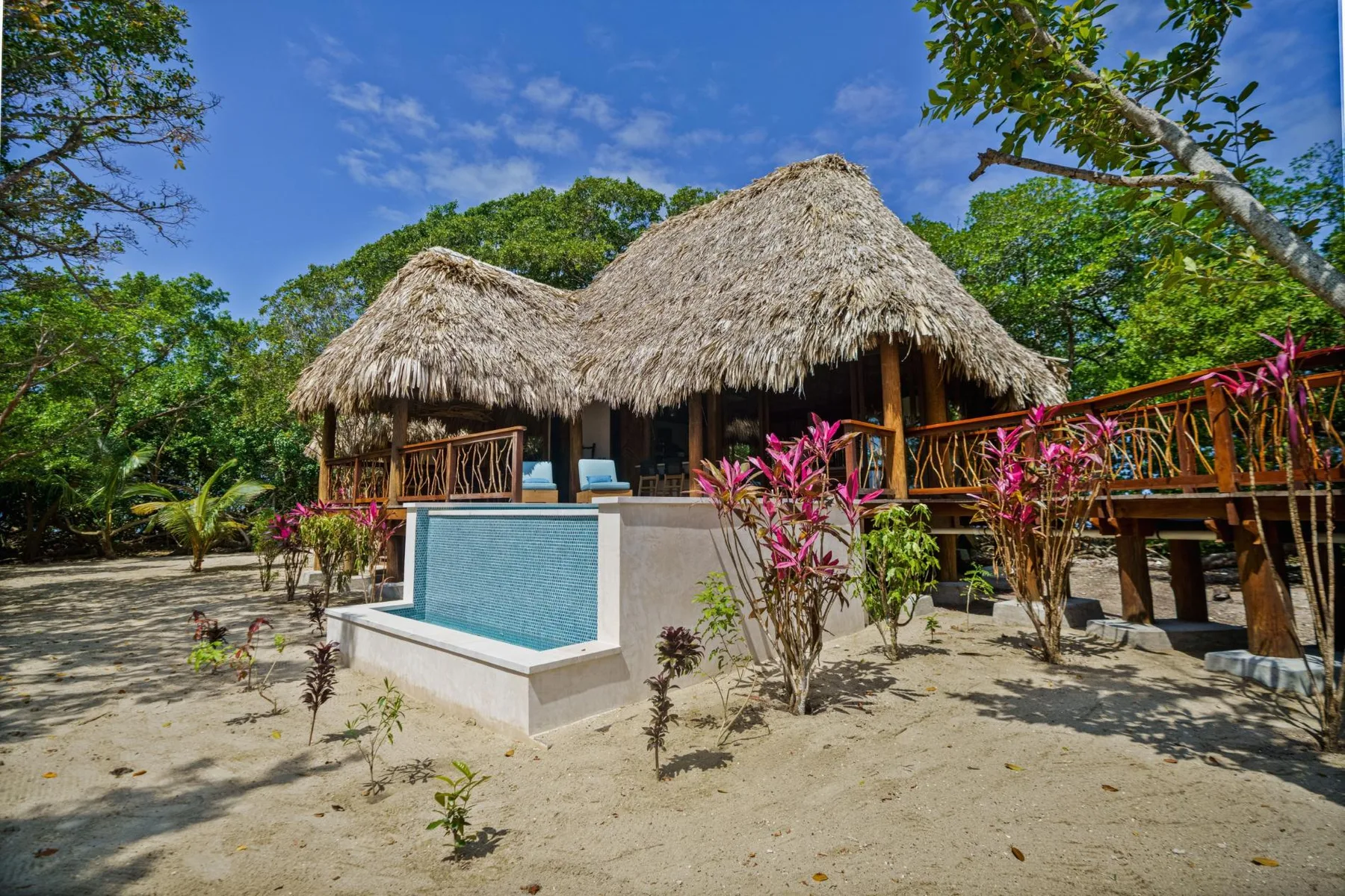 A thatched-roof tropical building stands behind a small tiled pool in a sandy garden.