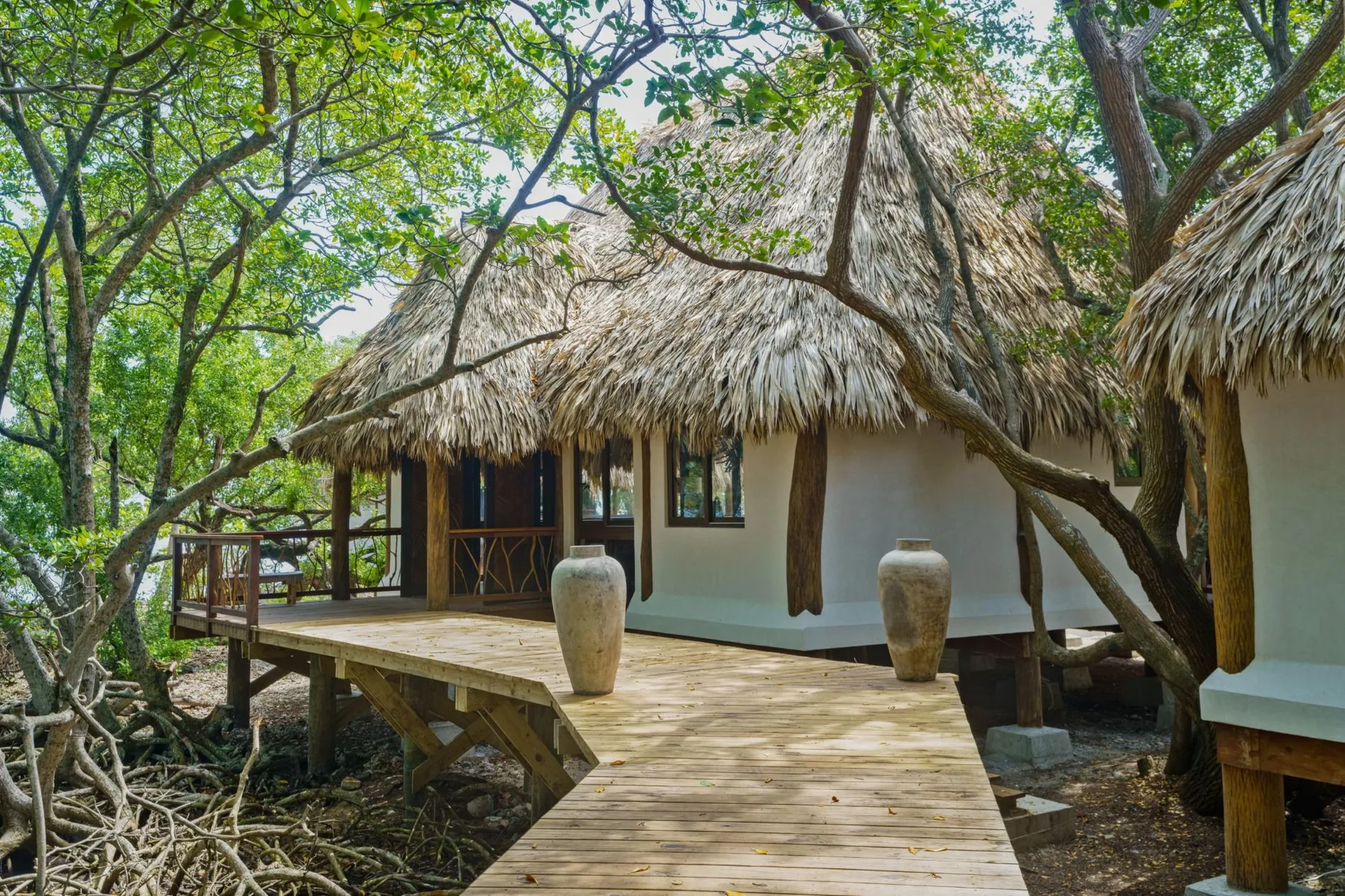 A thatched-roof hut stands beside a wooden walkway among leafy trees.