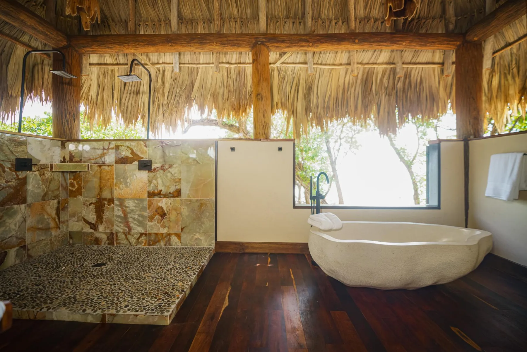 An open-air bathroom with a bathtub, shower, and broad windows beneath a thatched roof.