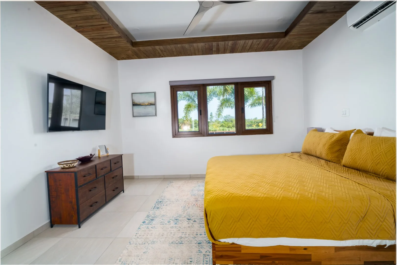 A bedroom with a yellow bedspread, wooden-framed window, and wall-mounted television.