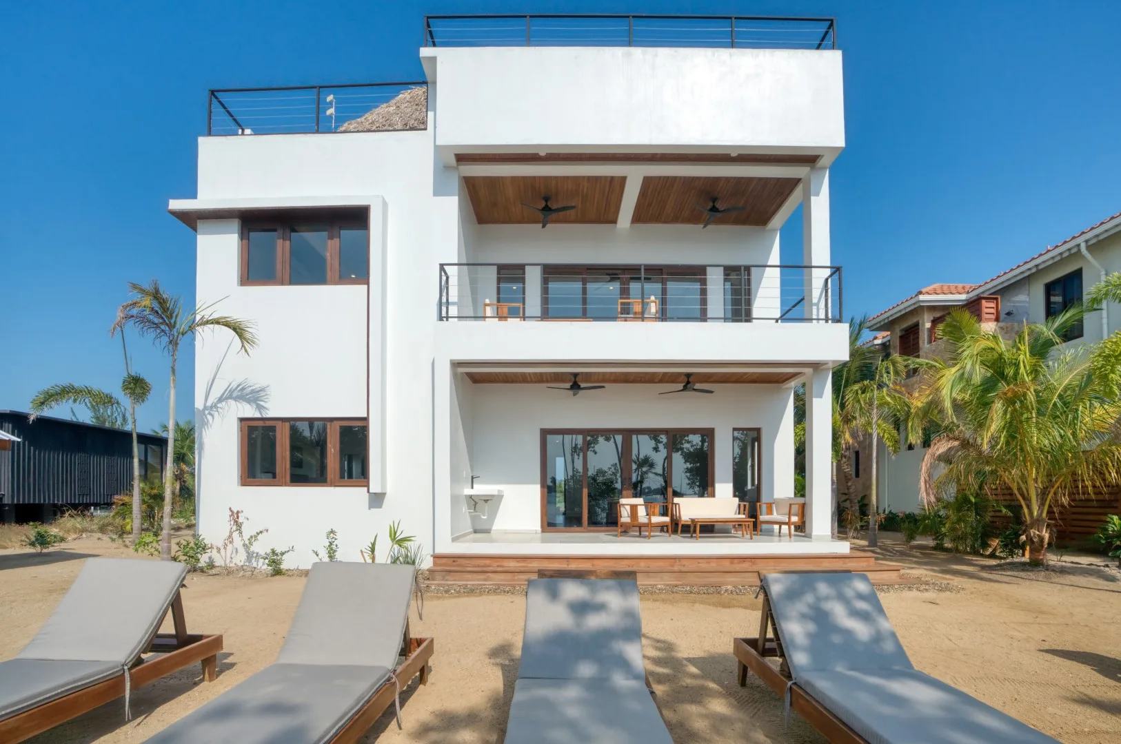 A modern white house with balconies and outdoor seating beside a sandy poolside area.