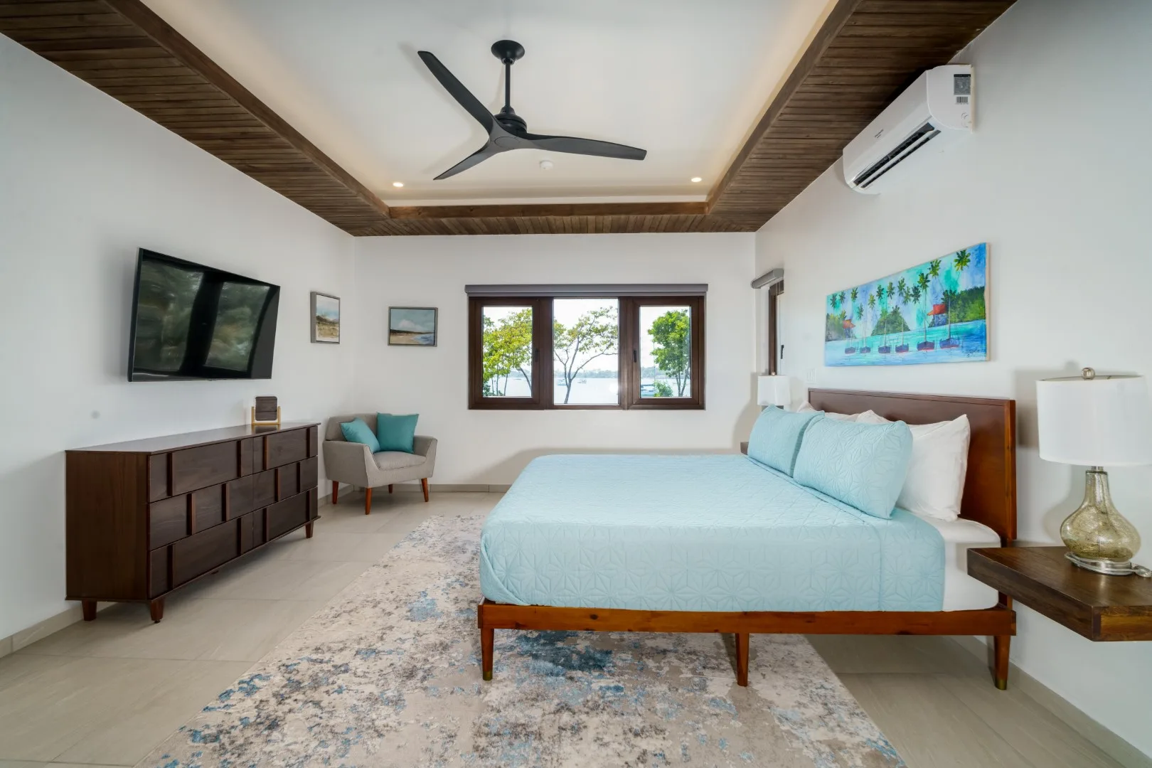 A bedroom with a bed, a television, and windows overlooking greenery.