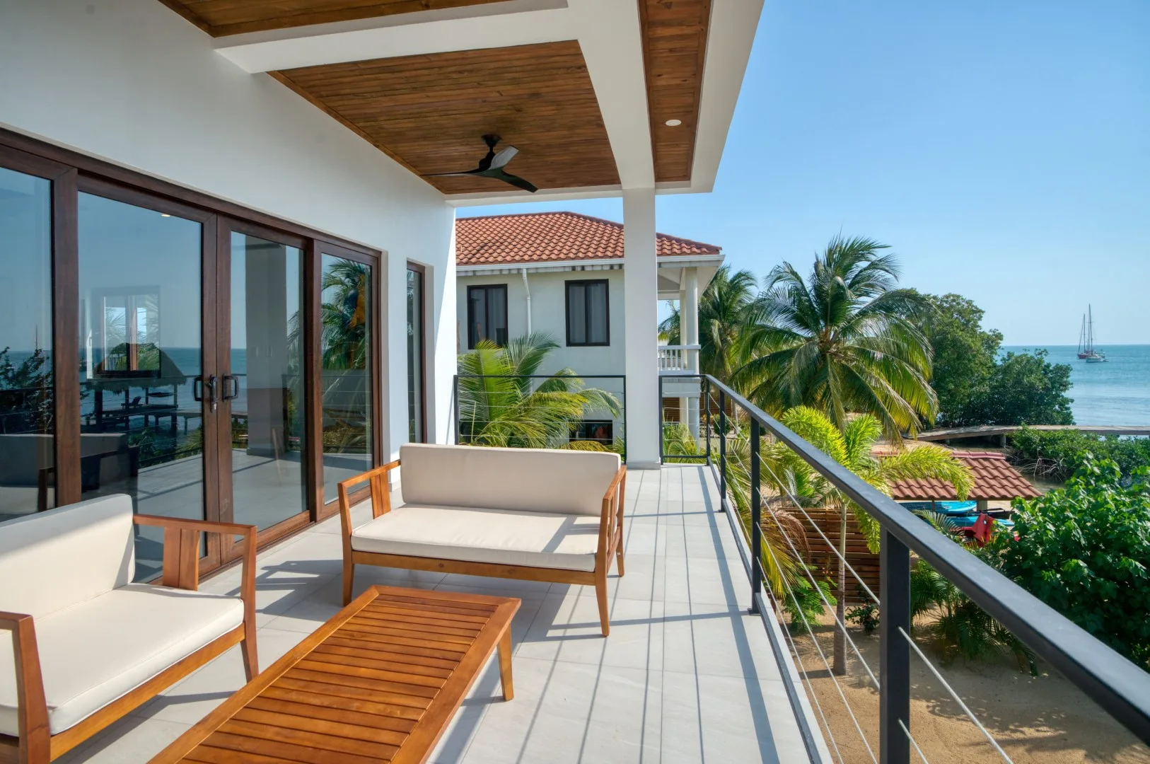 A covered balcony with outdoor seating overlooks palm trees and the sea.