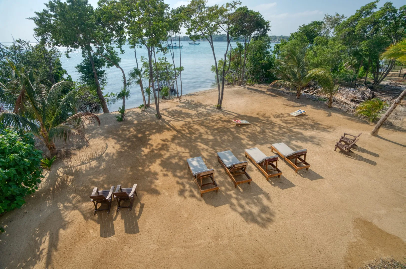 A sandy outdoor seating area beneath trees overlooks a calm body of water.