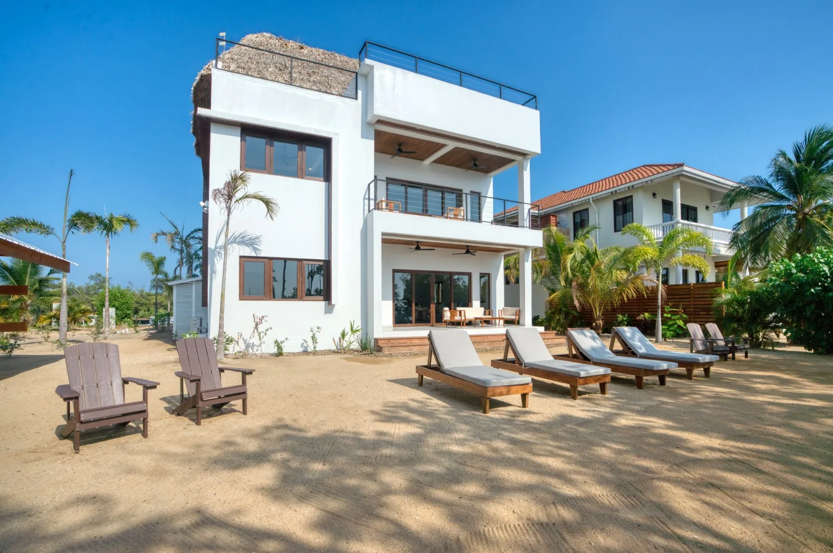 A modern white house stands beside a sandy patio with outdoor seating.
