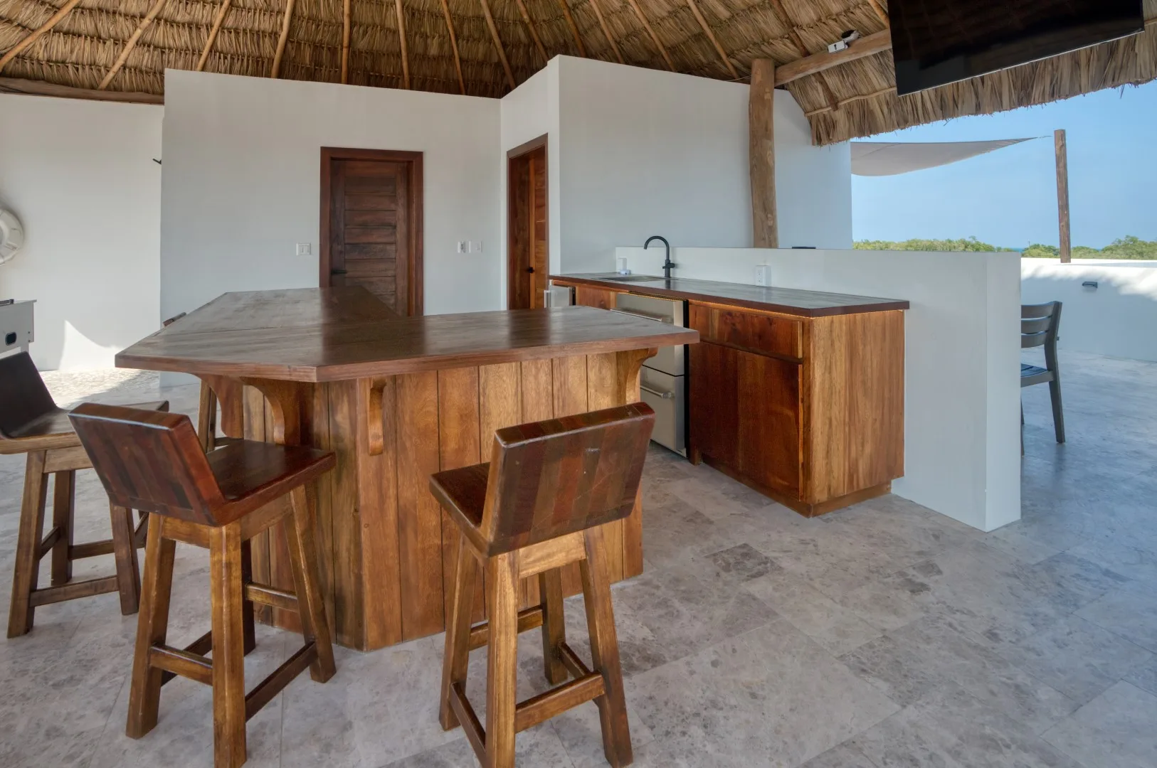An open-air kitchen and dining area with wooden seating beneath a thatched roof.