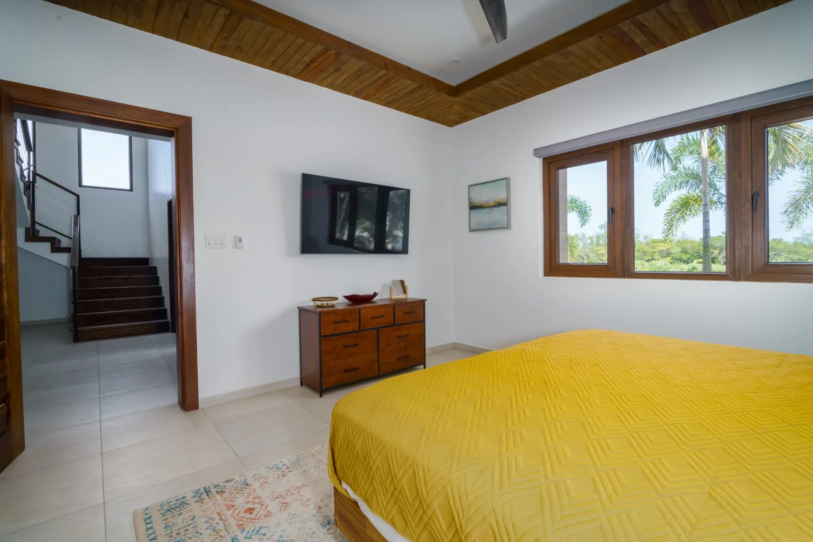 A bedroom with a yellow-covered bed and a window overlooking palm trees.