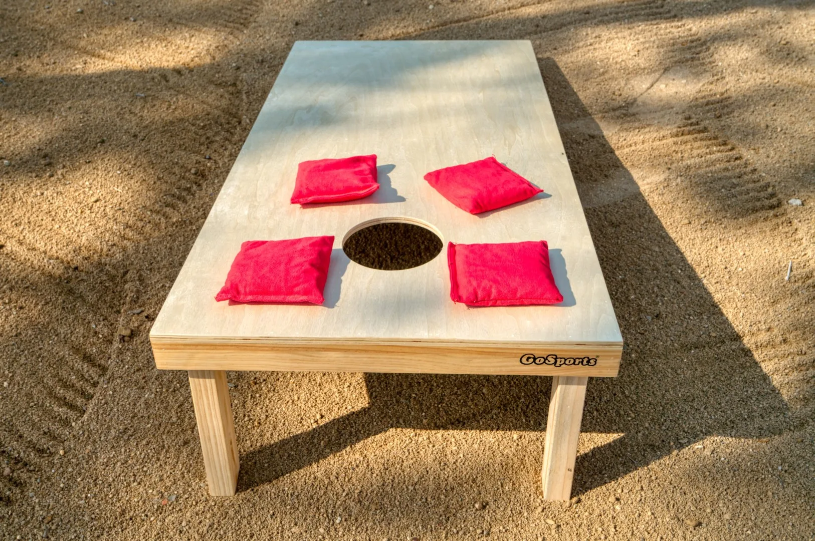 A cornhole game board rests on sandy ground with red beanbags.