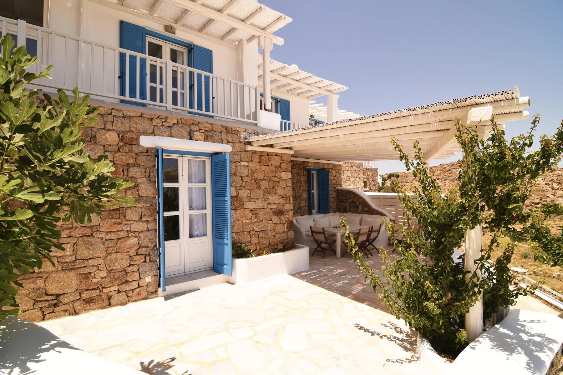 A sunlit stone house exterior with blue shutters and a shaded patio.