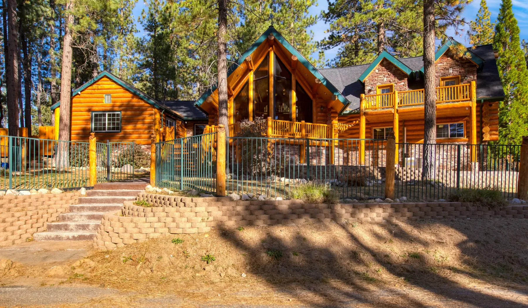 A large timber house stands among pine trees behind a metal fence.