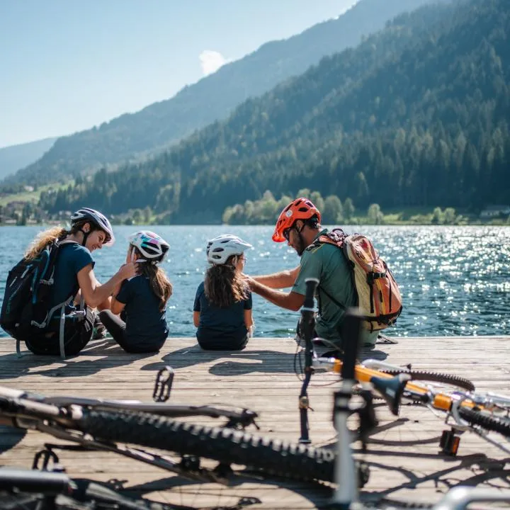 Cyclists rest beside bicycles on a lakeside dock, with wooded mountains across the water.