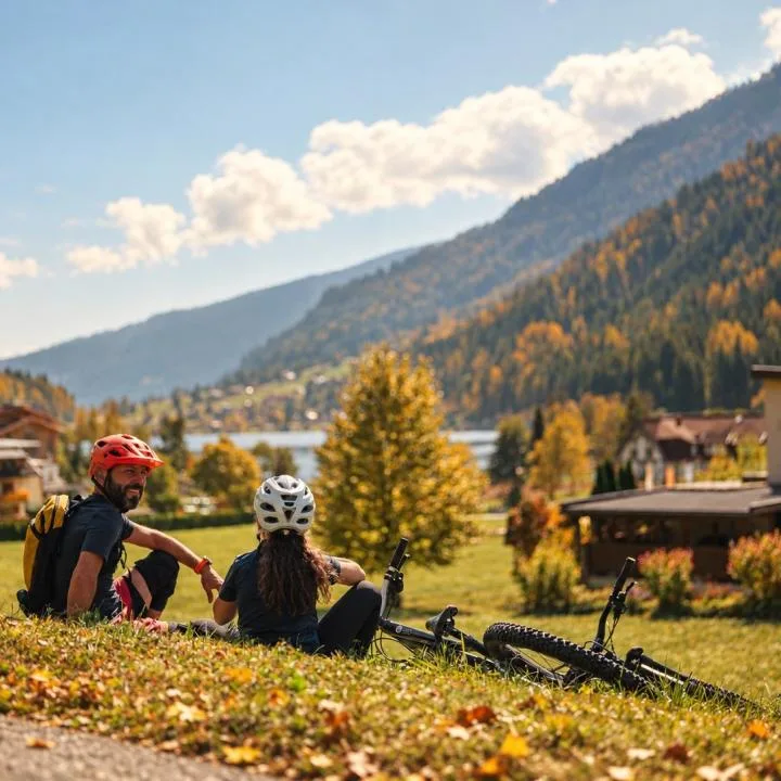 Two cyclists rest beside their bicycles on a grassy hillside overlooking a mountain village.