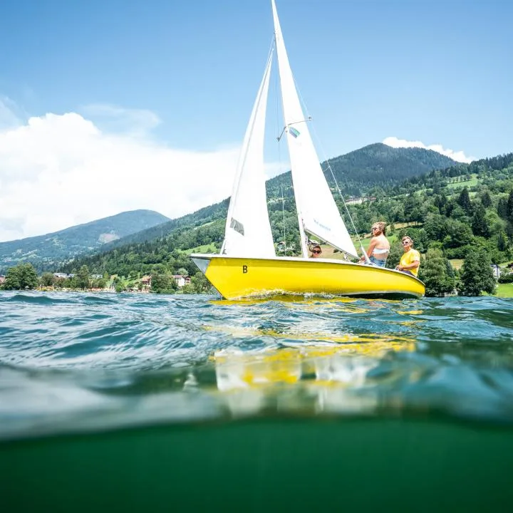 A yellow sailboat with people aboard glides across a lake beneath forested hills.