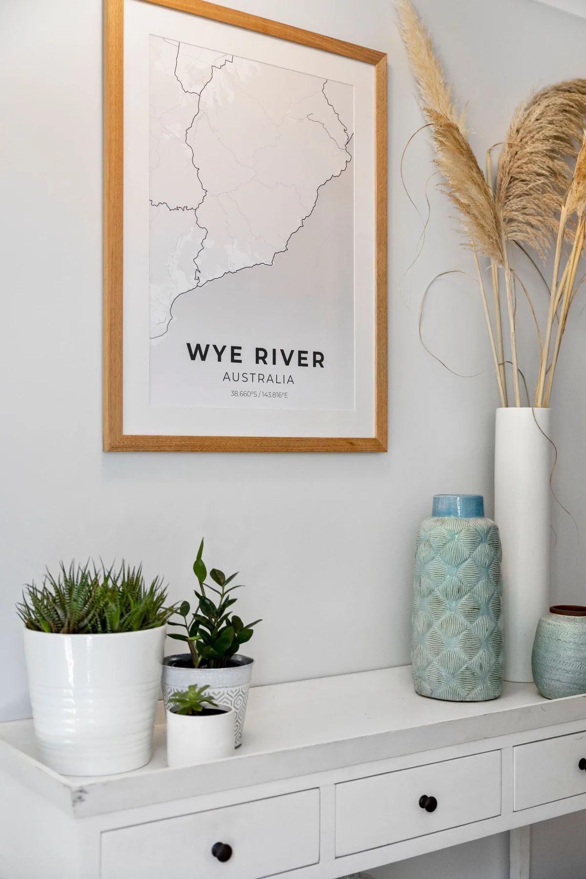 Una consola blanca exhibe plantas en macetas, un mapa enmarcado y un jarrón alto.