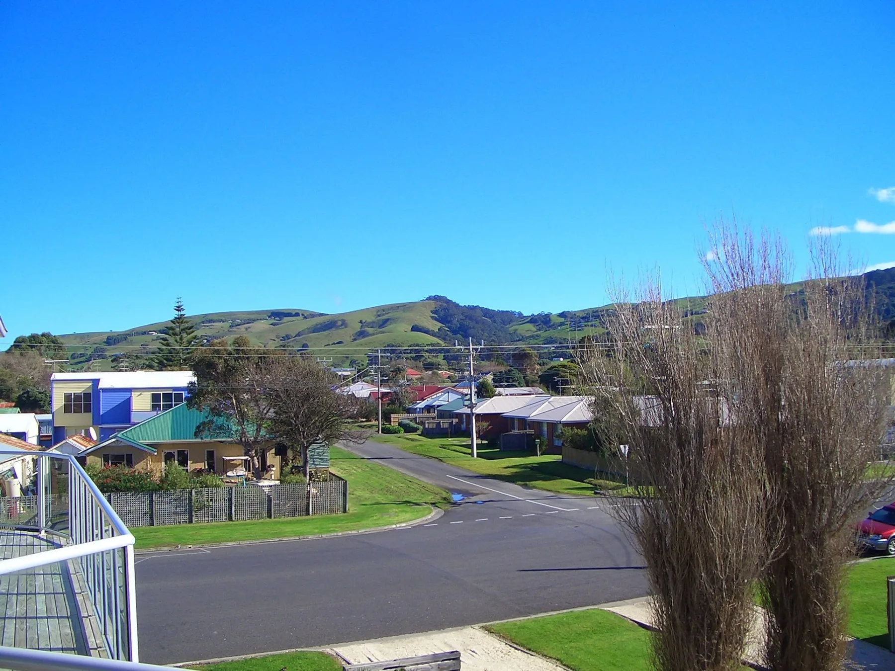 A residential street curves past houses and leafless trees, with green hills in the background.