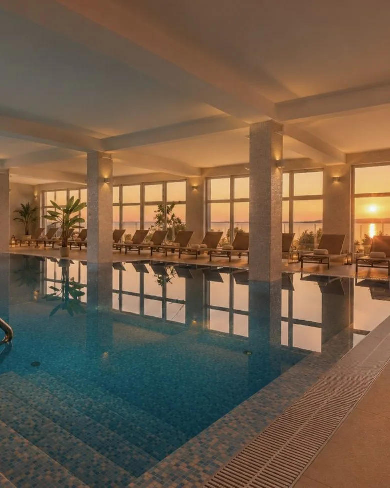 Representative hotel space: An indoor swimming pool reflects large windows and seating at sunset.