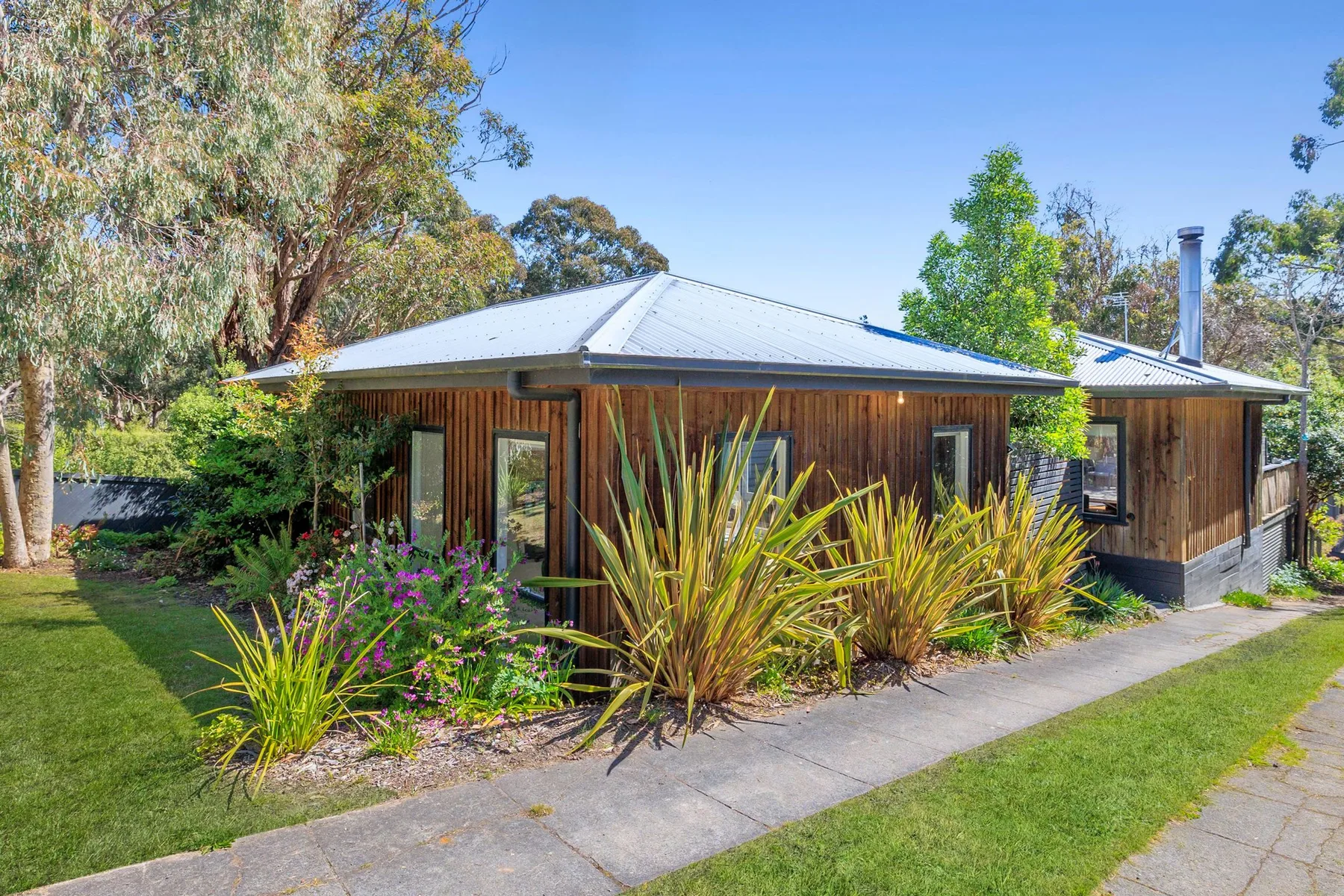 A wooden house sits among leafy garden plants beside a paved path.