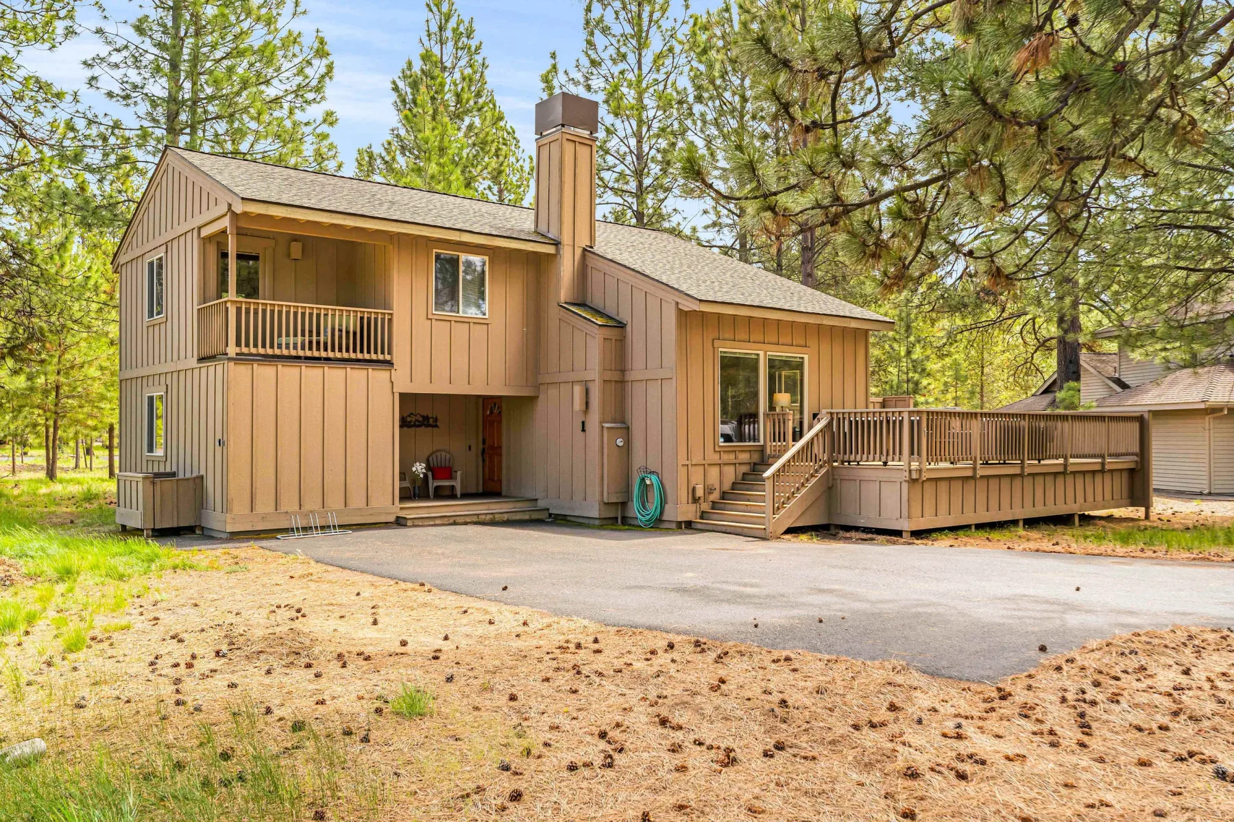 A two-story wood-sided house stands among pine trees beside a paved driveway.