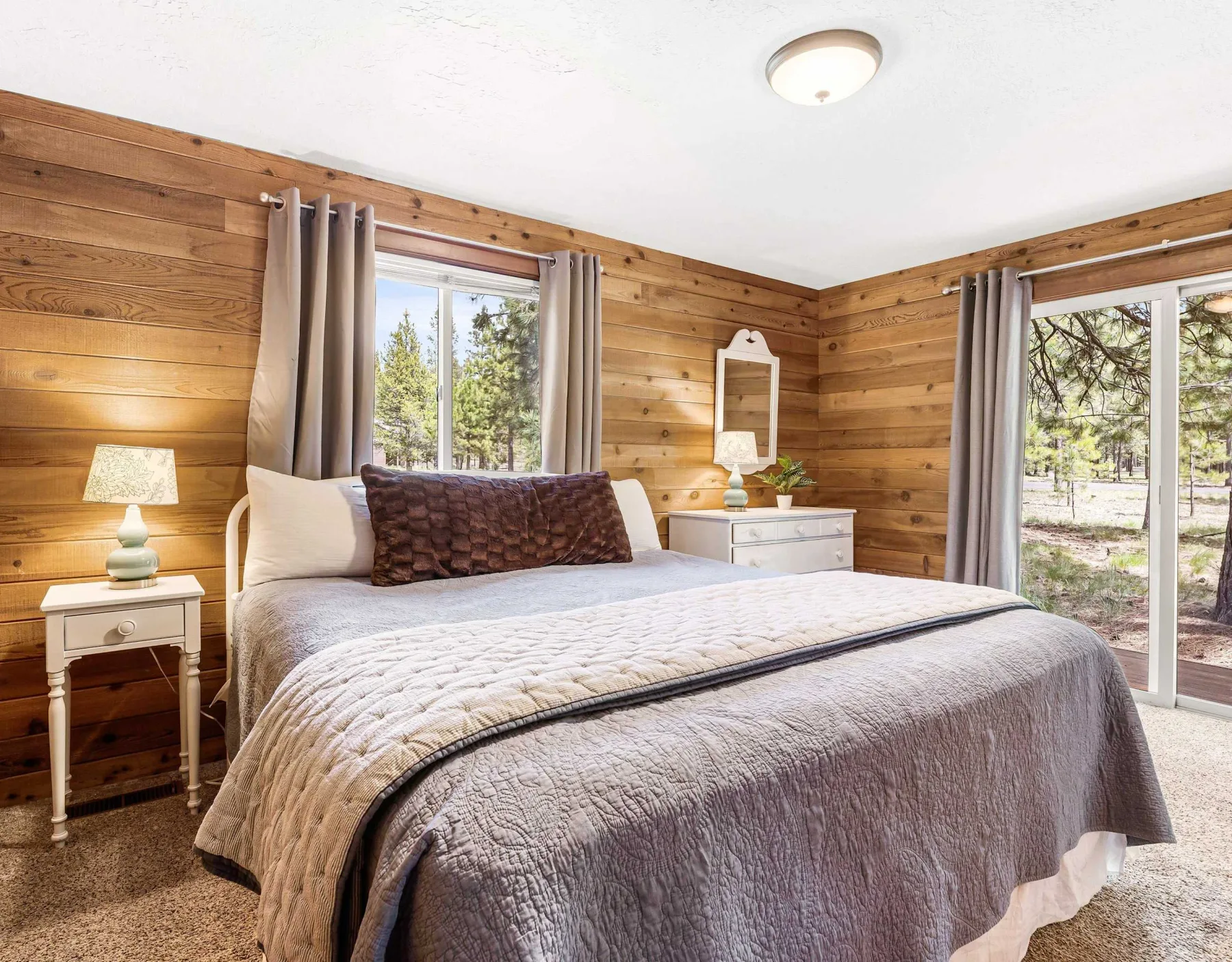 A bedroom with a neatly made bed and windows overlooking trees.