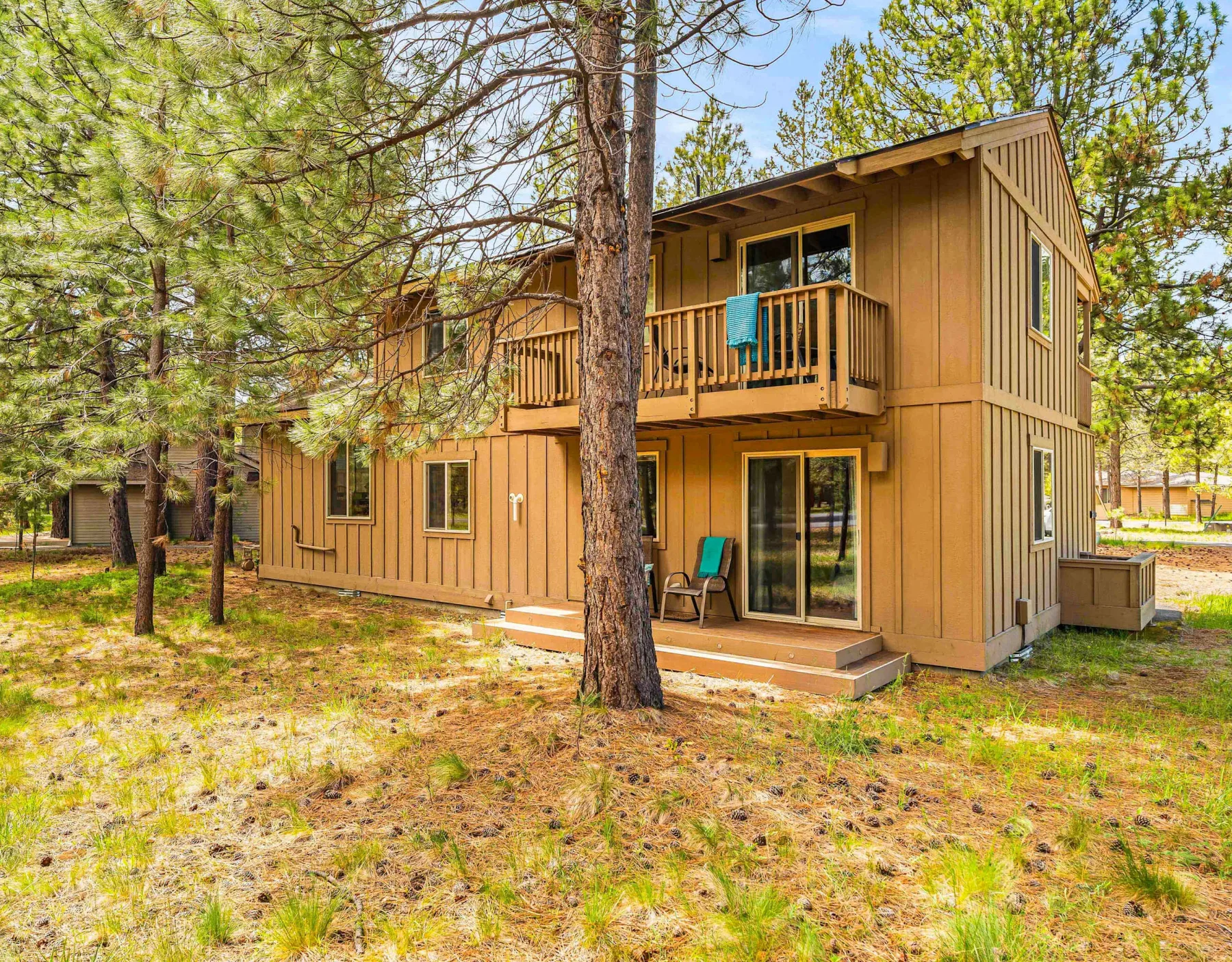 A two-story wooden cabin stands among pine trees, with a small balcony and ground-level patio.