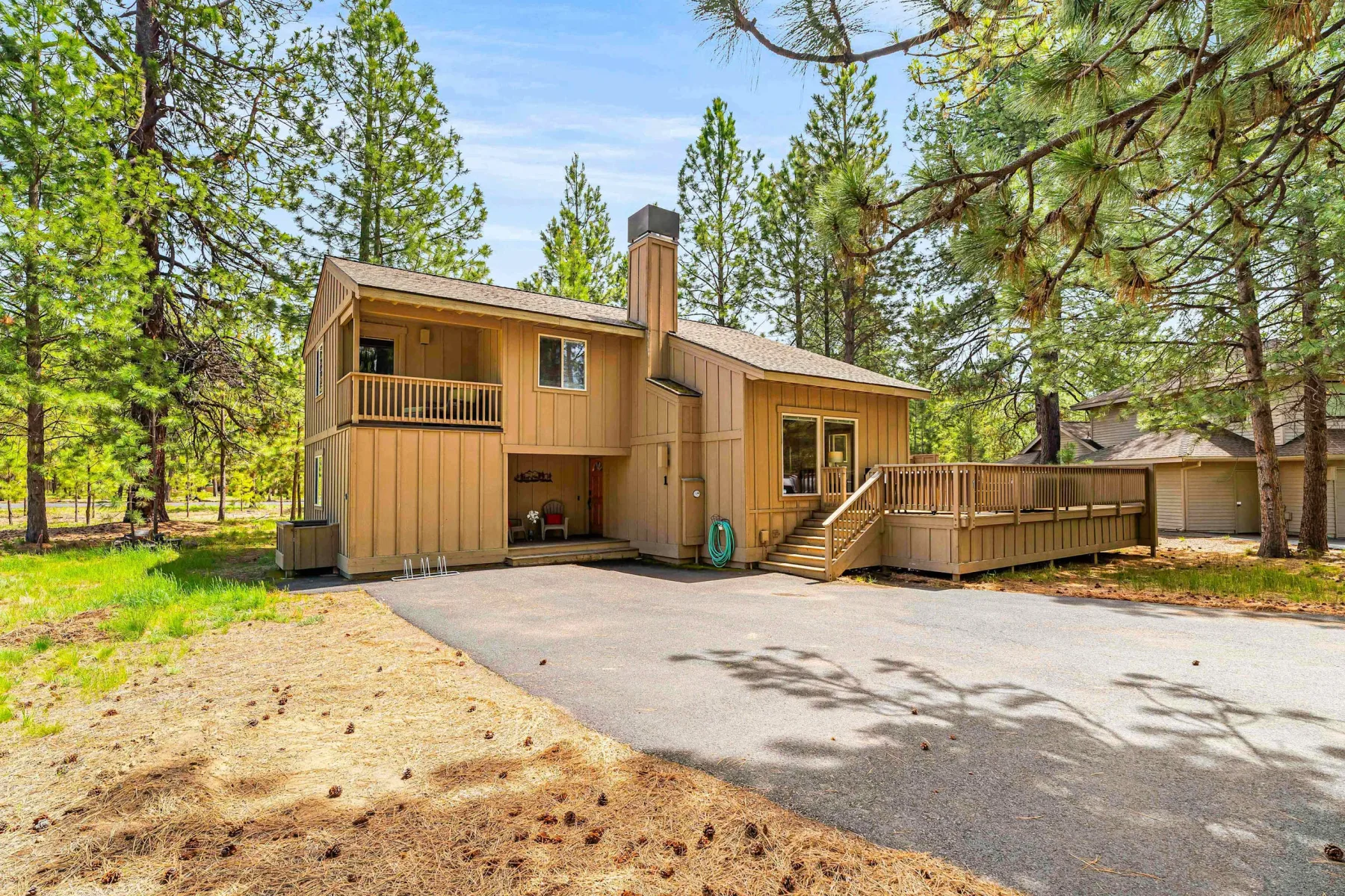 A wooden cabin with a raised deck and paved driveway among pine trees.