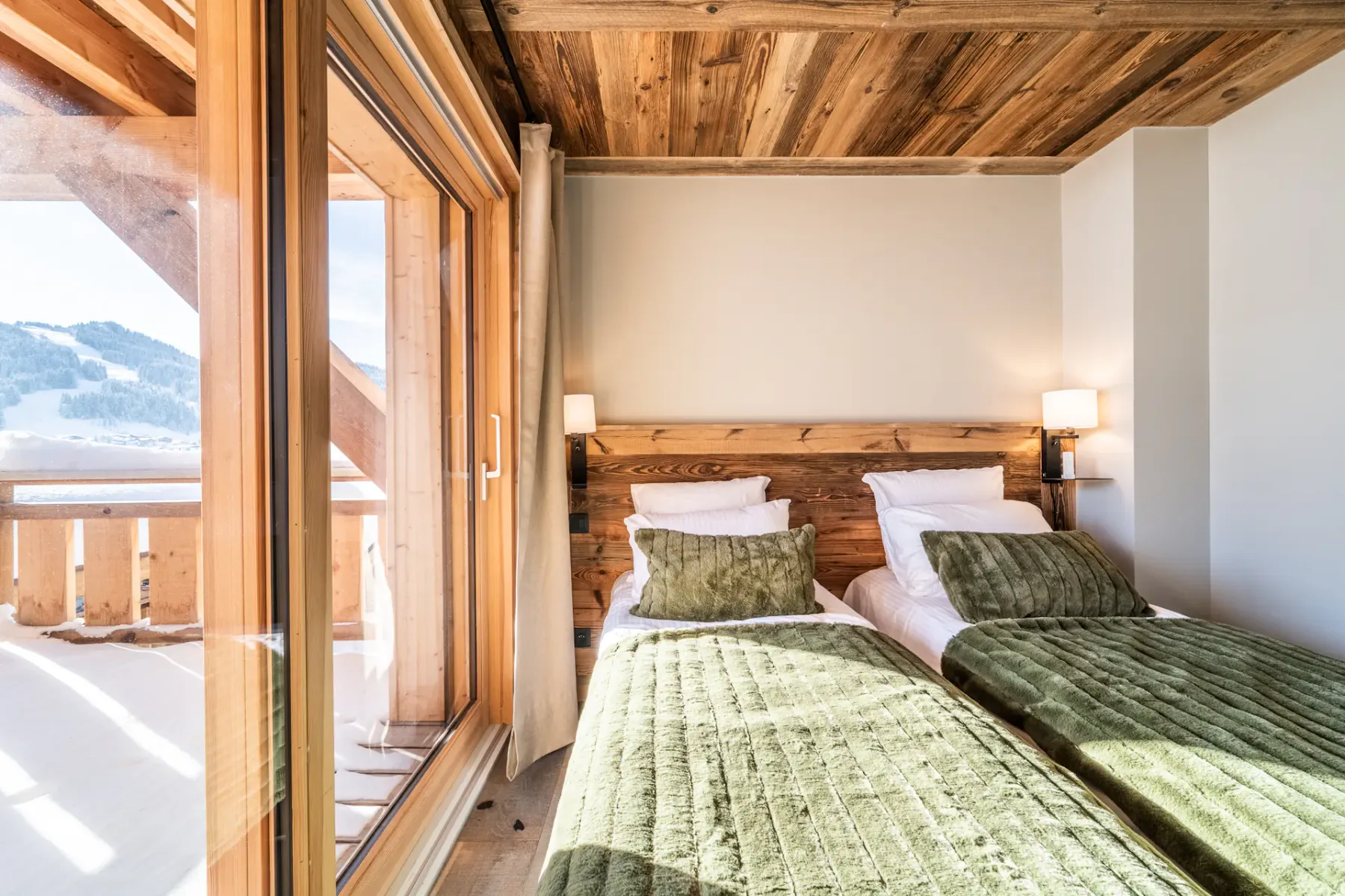 Un dormitorio con dos camas, ropa de cama verde y grandes ventanales con vistas a un paisaje nevado.