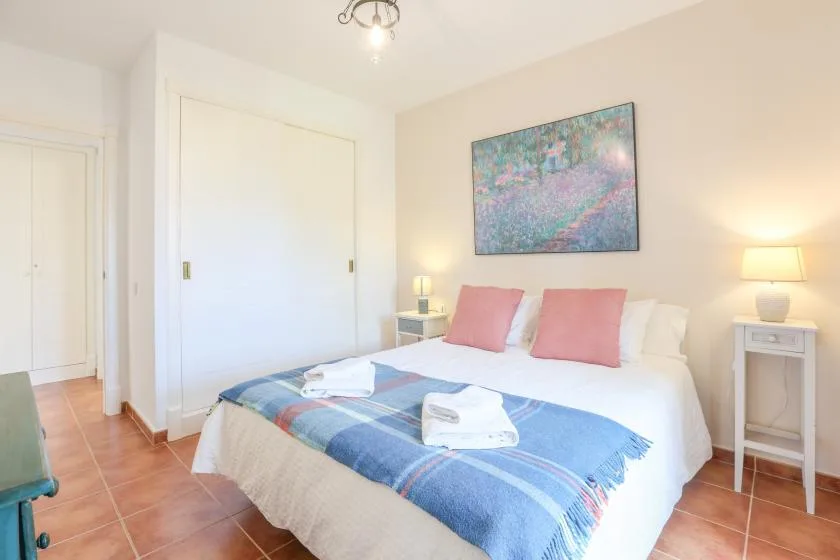 A bedroom with a bed covered by a blue blanket and a painting above it.
