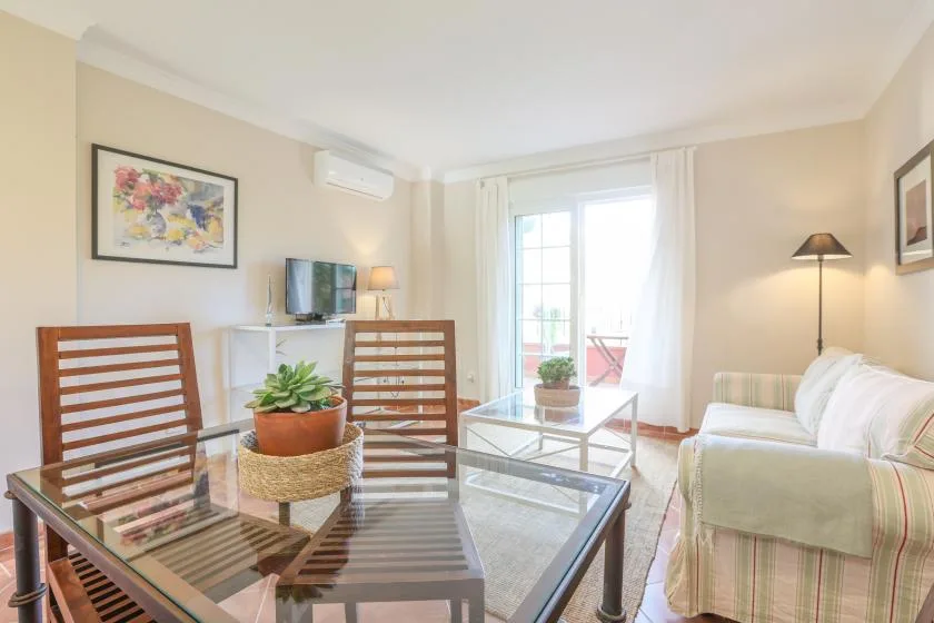 A bright living room with seating, a glass coffee table, and a large window.