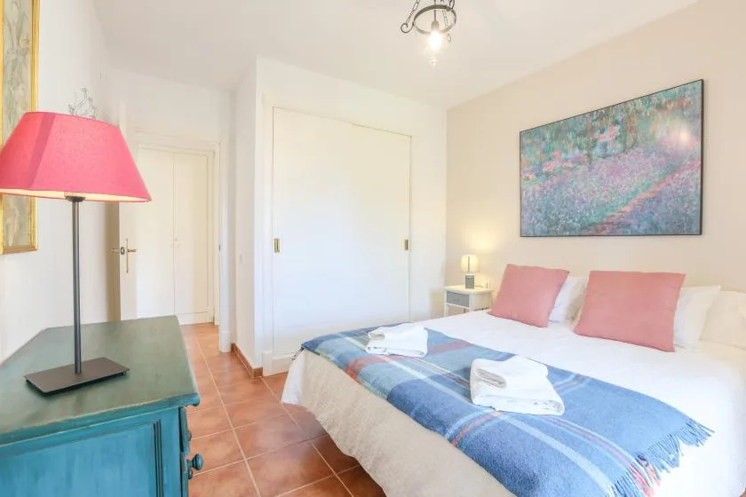 A bright bedroom with a bed, folded towels, and colorful artwork above it.