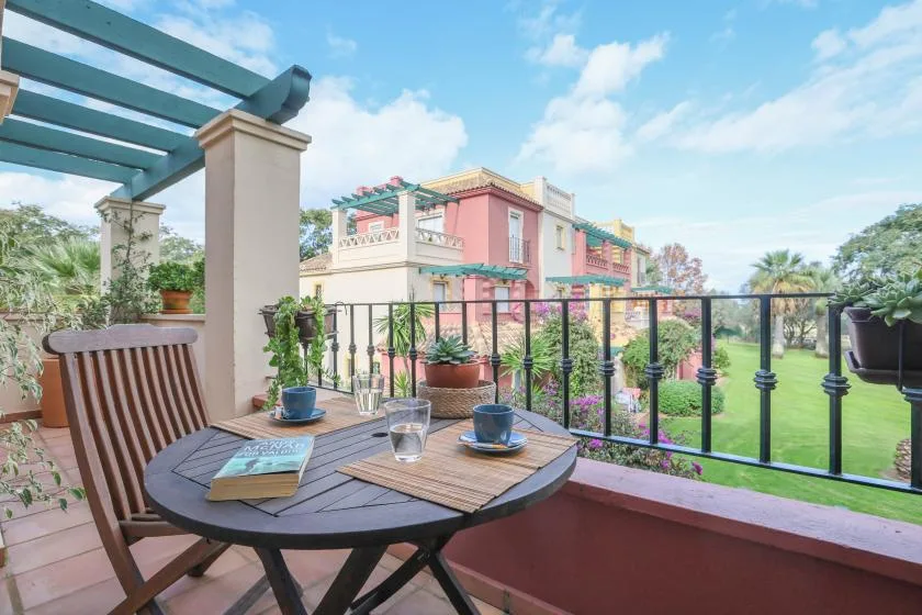 A patio table and seating overlook a garden and colorful building.